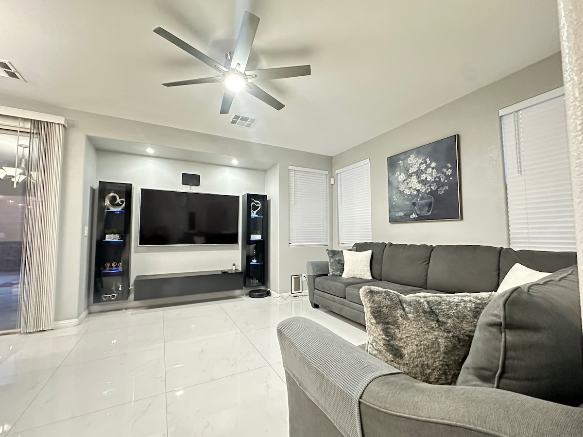 The living room is furnished with a sectional sofa and a comfortable armchair. A wall-mounted TV is positioned on a modern media console against a light-colored wall. Natural light enters through windows adorned with blinds, while a ceiling fan hangs over the seating area.