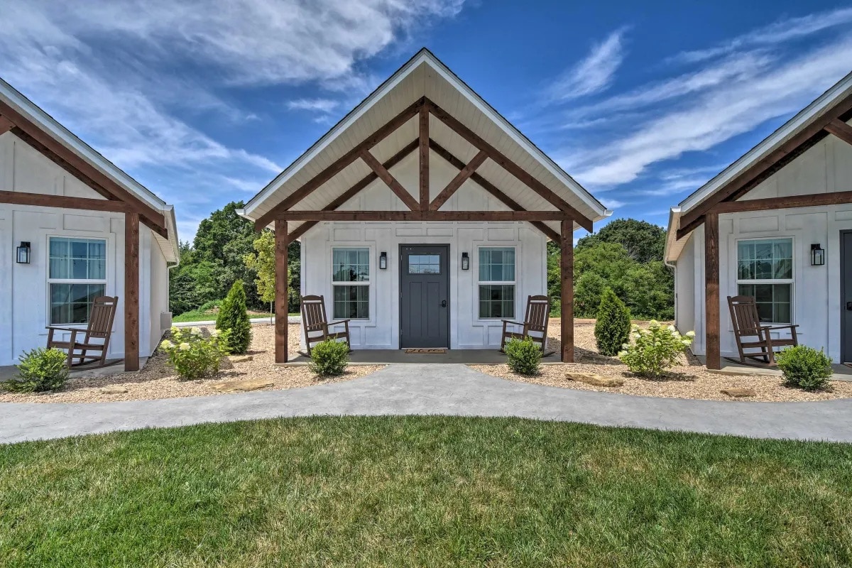 A charming cottage entrance is framed by two rocking chairs placed on a wide porch. Symmetrical landscaping is featured on both sides, with neatly trimmed shrubs and a gravel pathway leading to the front door. A clear sky and distant trees create a tranquil setting.