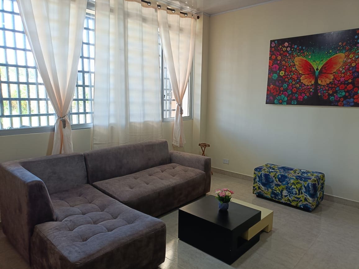 A comfortable living area features a large sectional sofa and a low coffee table. Bright curtains filter natural light from the windows, creating a relaxed atmosphere. A colorful butterfly painting adds a vibrant touch to the neutral walls, complemented by a small decorative plant on the table.