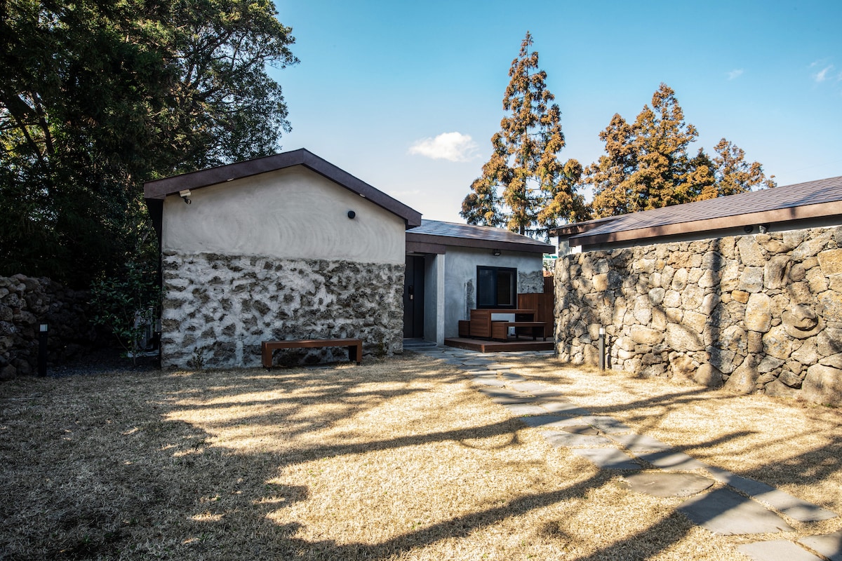 The exterior features a combination of rustic stone and modern architectural elements, with a pathway leading through a well-maintained grassy area. Surrounding tall trees provide a natural backdrop, enhancing the serene atmosphere of the space.