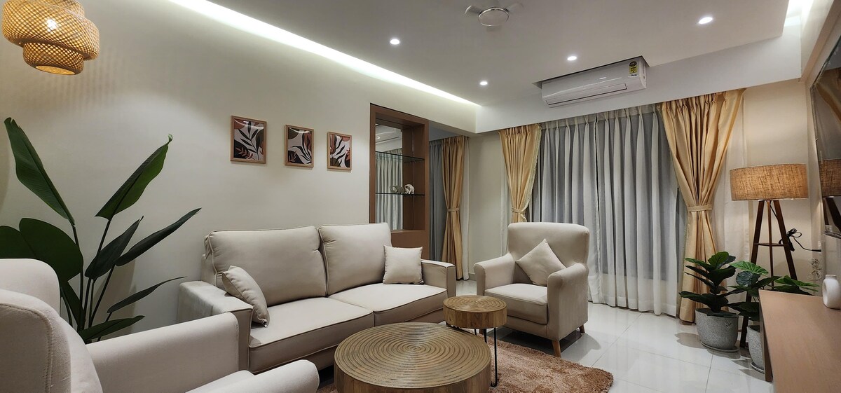 A welcoming living area is presented with soft upholstery and a round coffee table at its center. Natural light fills the space through large windows adorned with sheer curtains, while indoor plants add a touch of green. Decorative wall art accents the neutral color scheme.