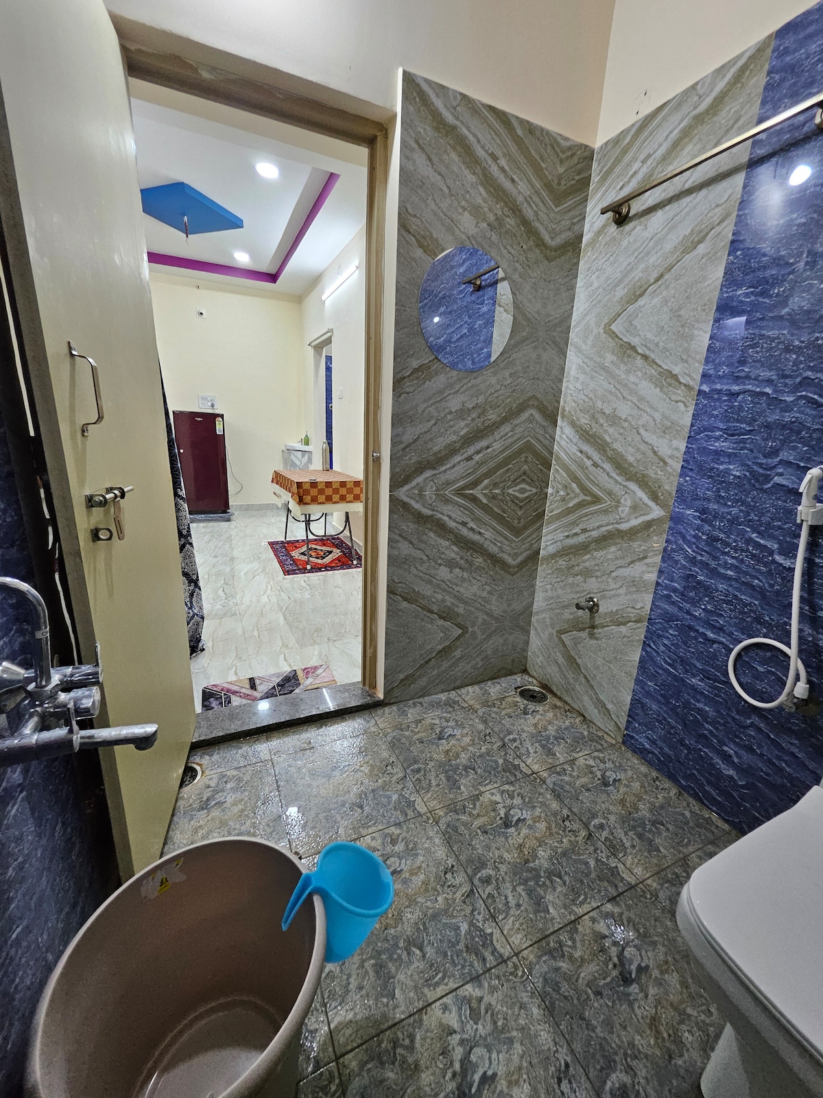A clean and modern bathroom is showcased with tiled walls in contrasting colors. A shower area is visible, along with a toilet and a bucket. The doorway leads to a well-lit interior space, suggesting convenience and accessibility.
