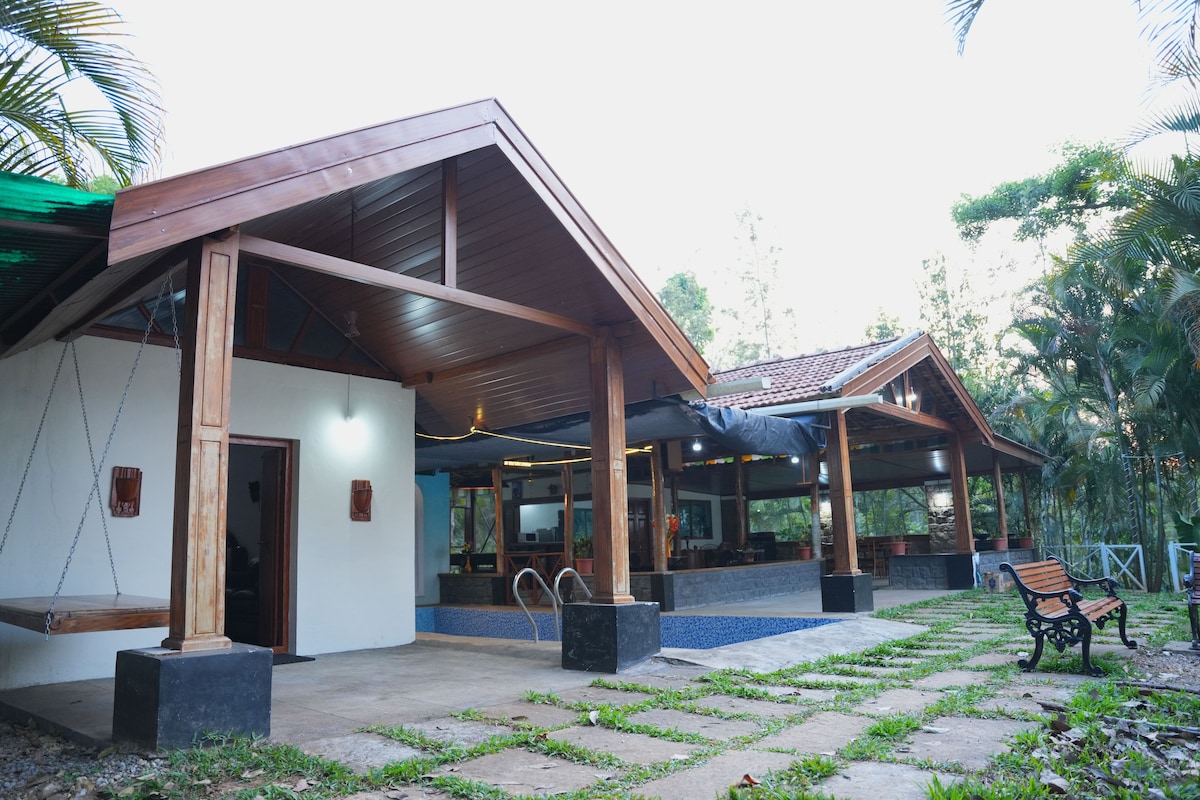 The image captures the exterior of a villa featuring a covered terrace area with wooden beams and an inviting private pool. A grassy area surrounds the pool, complemented by a wooden bench, with clear skies and lush greenery providing a serene backdrop.