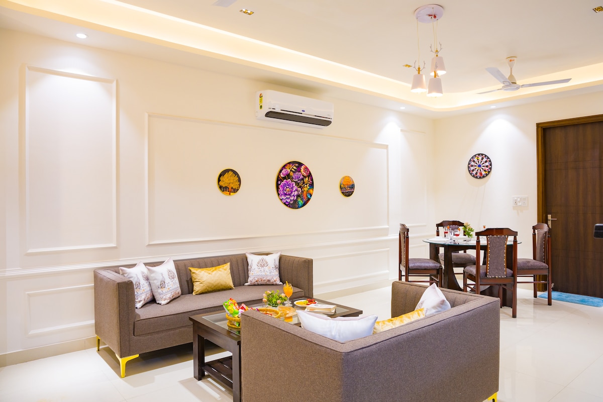 The living room features a light-colored wall with decorative plates and a sleek sofa set arranged around a central table. A dining area with a round table and four chairs is visible. Soft lighting enhances the space, while an air conditioning unit is mounted on the wall.