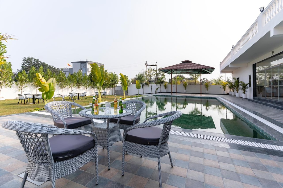 An outdoor seating area is arranged around a circular, stone table, featuring four wicker chairs with soft cushions. In the background, a rectangular pool reflects the blue sky and is complemented by a gazebo structure, surrounded by landscaped greenery.