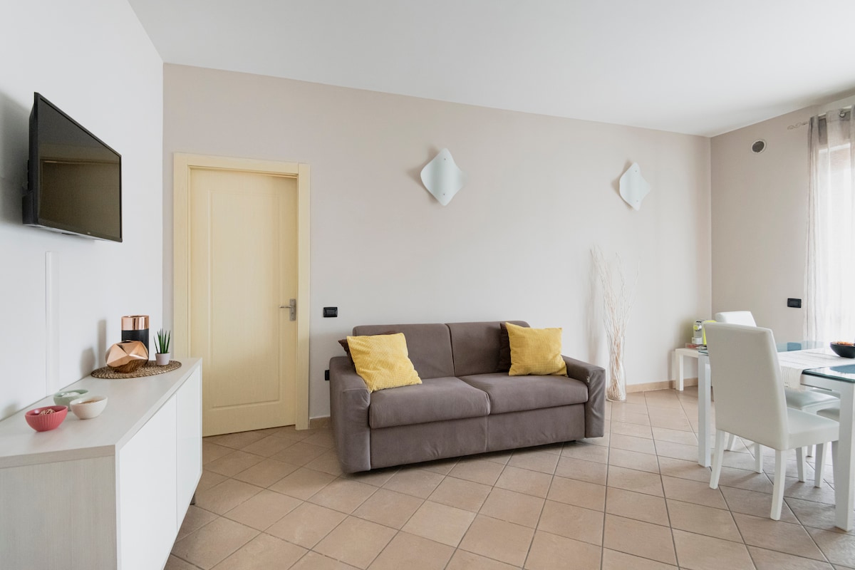A well-lit living area features a comfortable sofa adorned with yellow accent cushions. A flat-screen TV is mounted on the wall, while a dining table with light chairs is set nearby. Soft natural light filters through the window, complementing the neutral-toned walls.