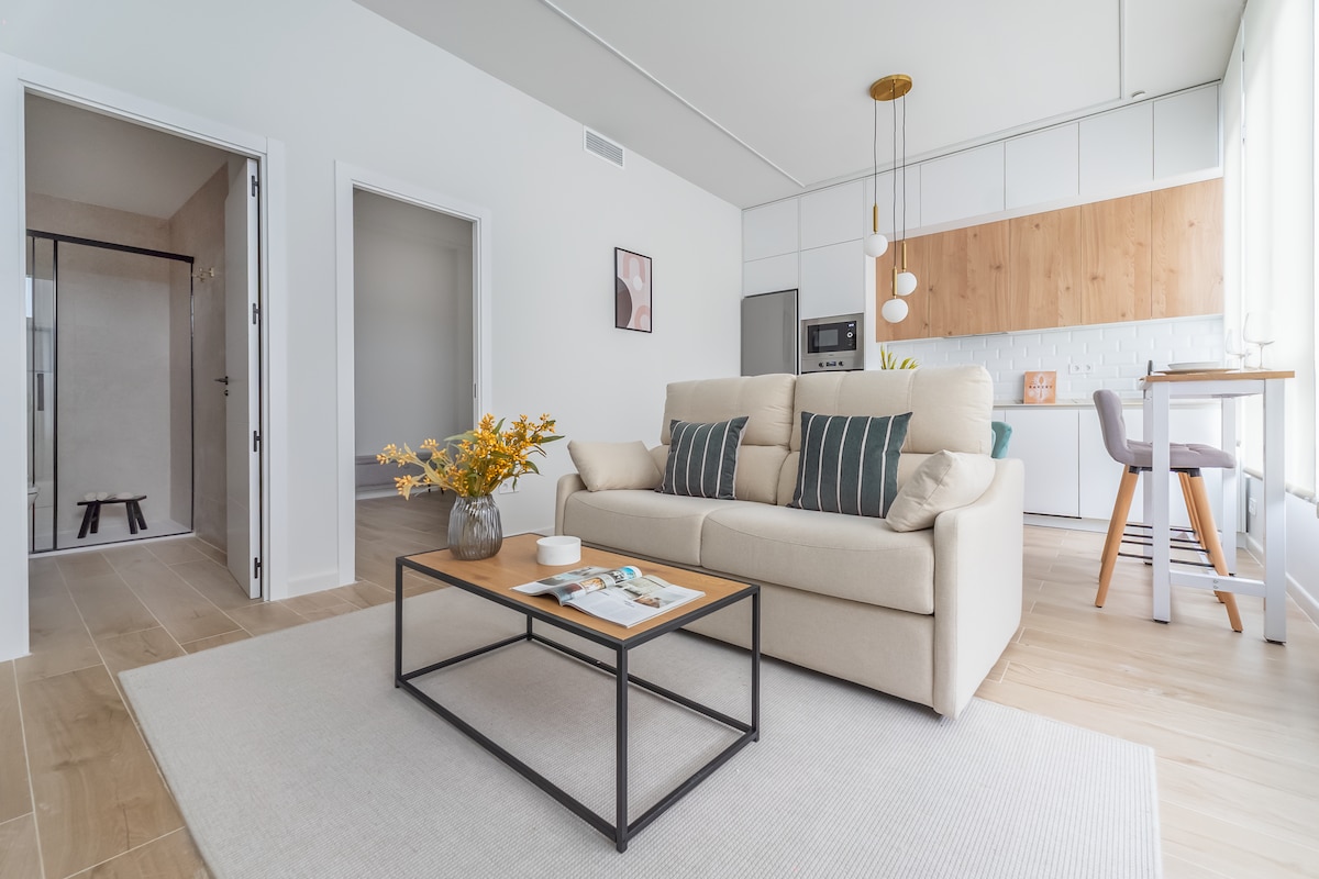 A modern living space features a comfortable sofa with decorative cushions, positioned in front of a stylish coffee table. A light, neutral colour palette is complemented by a minimalist kitchen in the background, showcasing wood accents and contemporary furnishings. Natural light streams in through large windows.