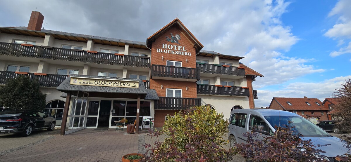 A welcoming hotel exterior is displayed, featuring three levels with balconies and a covered entrance. The building is flanked by greenery and a paved area where parked vehicles are visible. The sky is partly cloudy, providing a soft light to the scene.