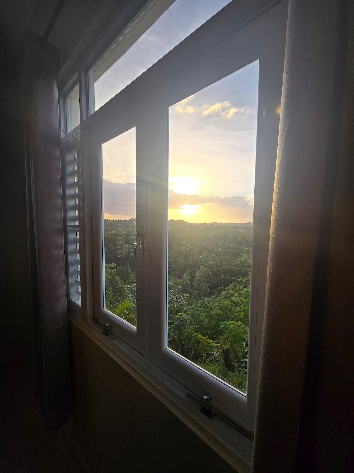 A large window frames a scenic view of the landscape, where the setting sun casts warm light over lush greenery. Soft curtains partially cover the window, creating a serene atmosphere within the room.