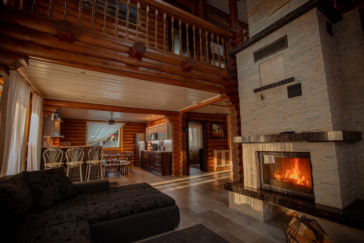 The spacious living area features a stone fireplace, providing warmth and comfort. A large sofa is positioned nearby, while a dining table and chairs are visible in the background. Warm light filters through expansive windows, enhancing the natural wood elements of the space.