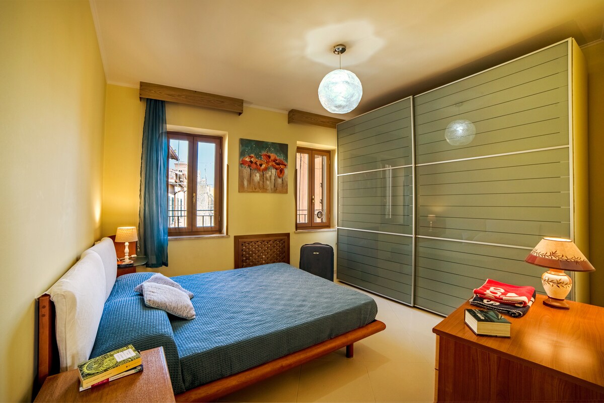 A bright bedroom features a spacious bed with a blue coverlet, accompanied by wood furnishings. Two windows allow natural light to fill the room, revealing modern decor including a wardrobe and bedside table. A soft lamp and an artwork piece are thoughtfully placed within the space.