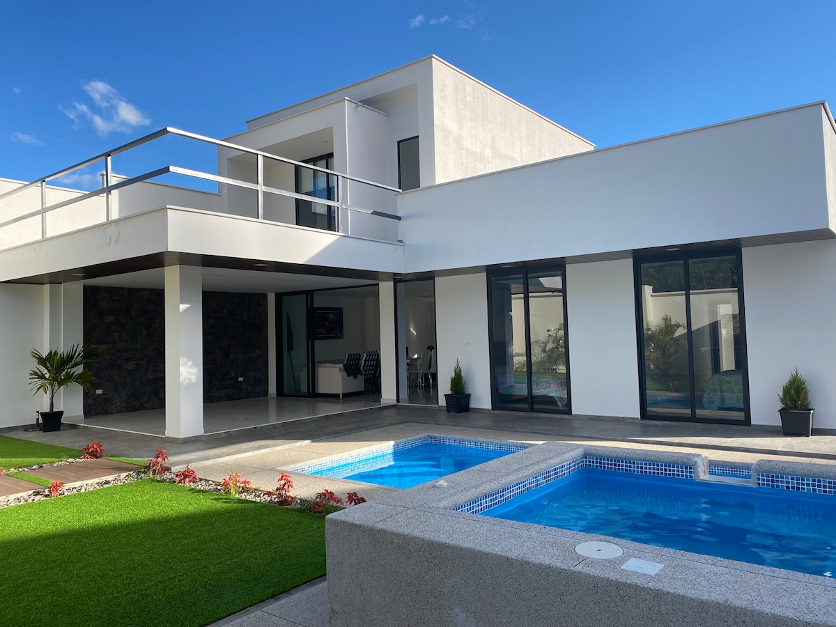 A modern home is presented with a sleek exterior, featuring large glass windows and a spacious balcony. A private pool is located in the foreground, surrounded by a landscaped area with green grass and decorative plants.