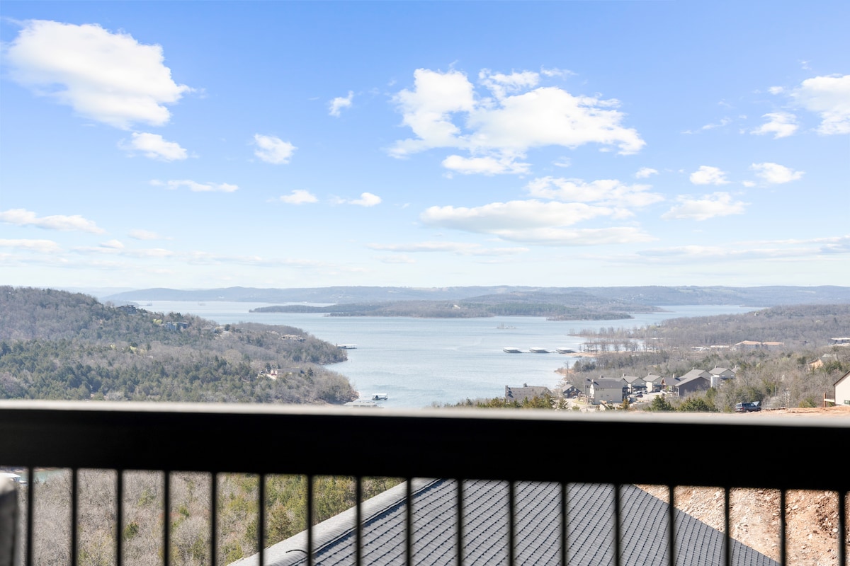 A scenic view of a tranquil lake is captured from a balcony, showcasing lush green hills on the horizon under a clear blue sky. Soft clouds drift above, accentuating the natural beauty of the landscape.