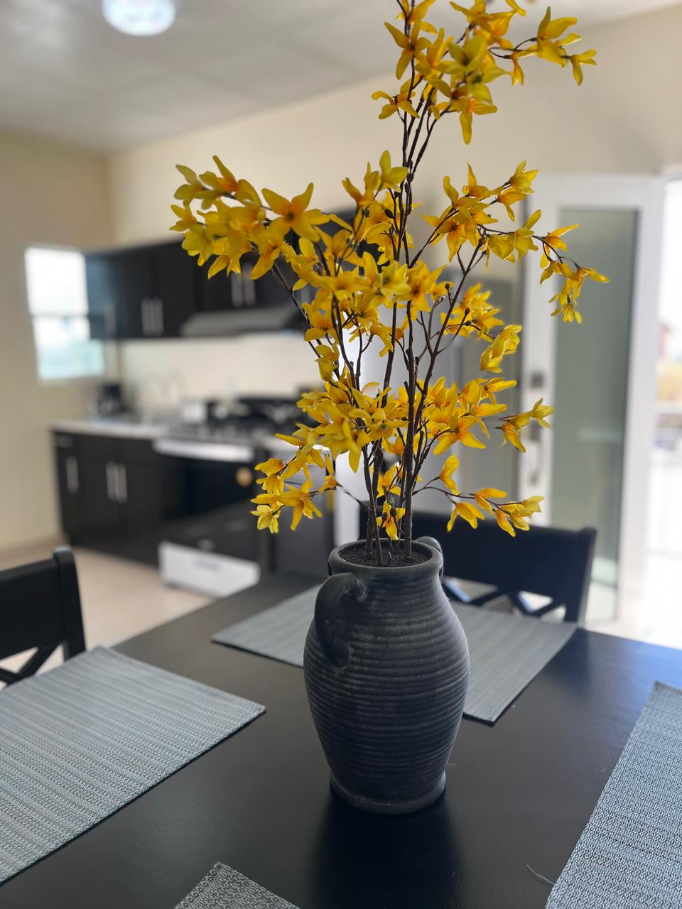 A dining table is set with gray placemats, complemented by a dark vase filled with yellow flowers. The background features a modern kitchen with dark cabinetry and a window allowing natural light to illuminate the space.