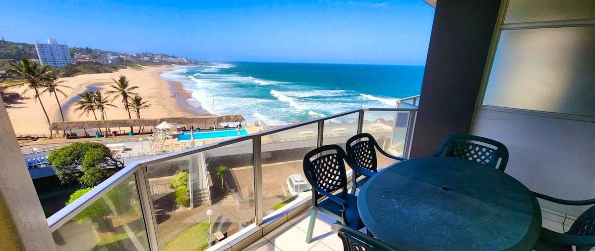 A balcony area is visible, featuring a round table surrounded by several chairs. The ocean and sandy beach can be seen in the background, with gentle waves rolling towards the shore. Palm trees are lined along the beach, contributing to the coastal view.