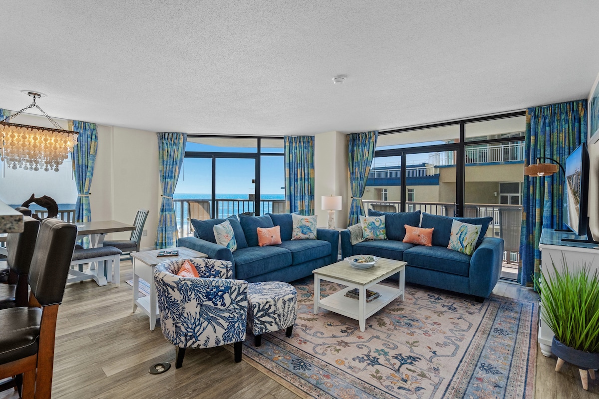A spacious living area is showcased, featuring a blue sectional sofa adorned with colorful throw pillows. Large windows allow natural light to fill the room, with ocean views visible in the background. A decorative area rug complements the seating arrangement, and a dining table is seen in the distance.