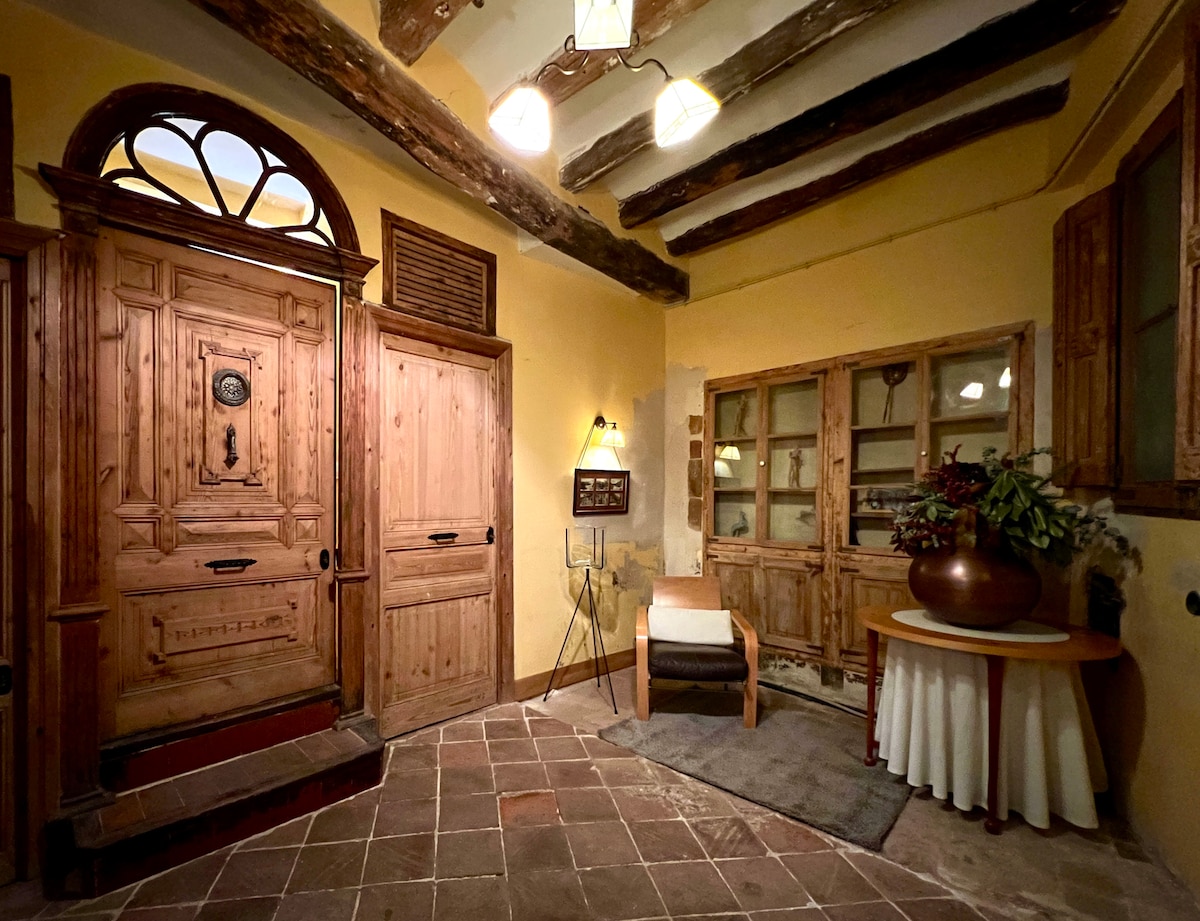 The entrance features a wooden door with intricate detailing, framed by exposed wooden beams and warm yellow walls. A small armchair is placed next to a round table with a vase, while a light fixture adds illumination to the space.