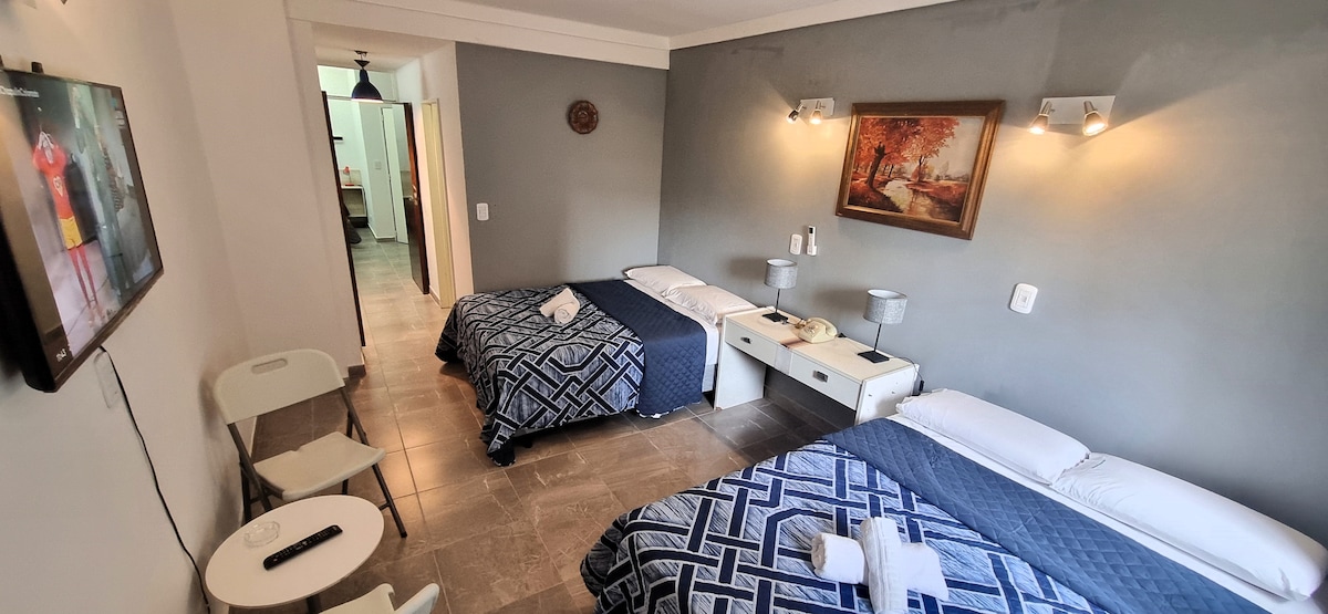 A spacious room is furnished with two beds, each topped with patterned blue bedding. A white side table sits between the beds, flanked by modern lamps. A television is mounted on the wall, and a painting adds a touch of color. A doorway leads to another area of the apartment.