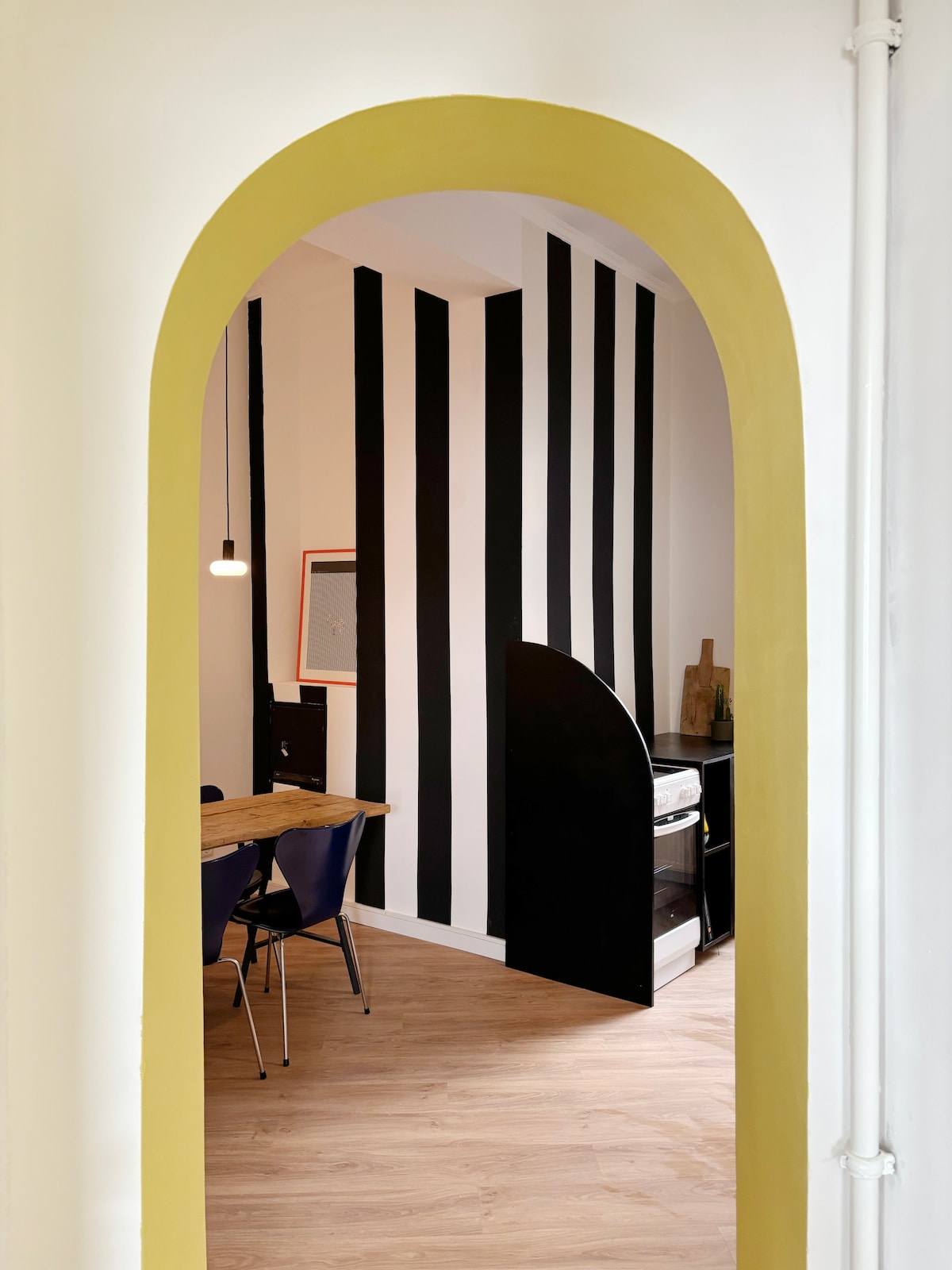 A dining area is visible through an arched entryway, featuring a wooden table surrounded by blue chairs. Bold black and white striped walls add a modern touch, while a sleek, dark partition offers separation from the adjoining space. Warm lighting enhances the inviting ambiance.