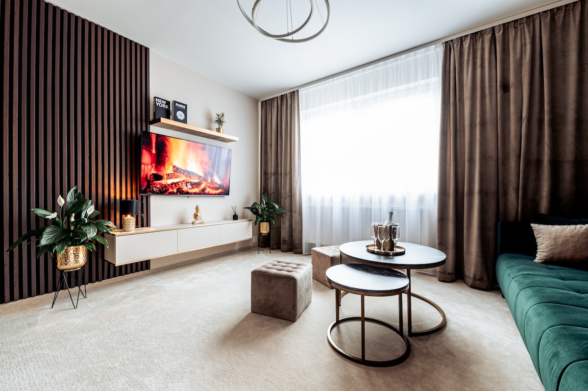 A modern living room features a large television mounted on a wooden accent wall. A plush sofa and a pair of circular coffee tables are arranged on a light-colored carpet. Natural light filters through a sheer curtain, and green potted plants add a touch of greenery.