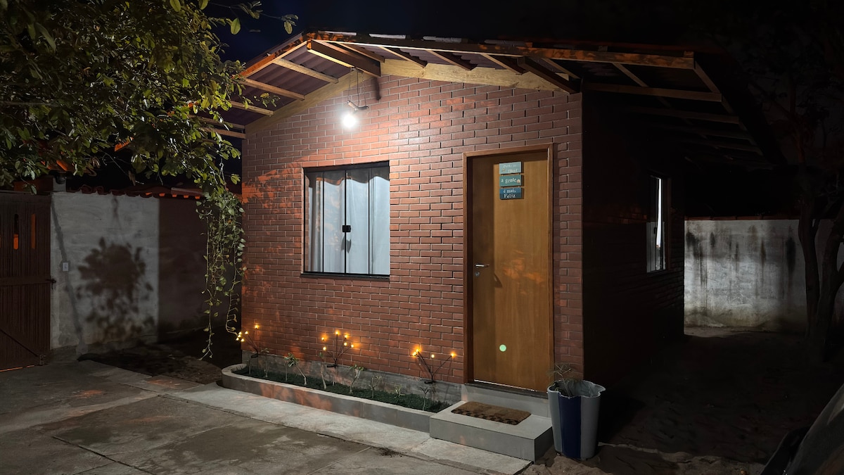 A cozy brick chalet exterior is illuminated by warm lighting in the evening. The space features a wooden door and window. A small garden area with lights and potted plants is visible. The surrounding area is paved and bordered by walls for privacy.