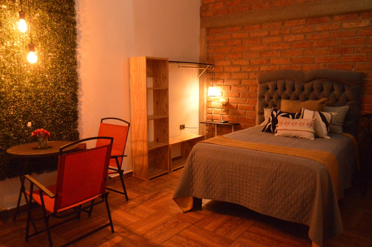 A sleeping area features a spacious bed with a grey quilt and decorative pillows. A cozy seating arrangement includes two red chairs and a small round table. A wooden shelf and a warm light fixture enhance the inviting ambiance, while a textured green wall adds visual interest.