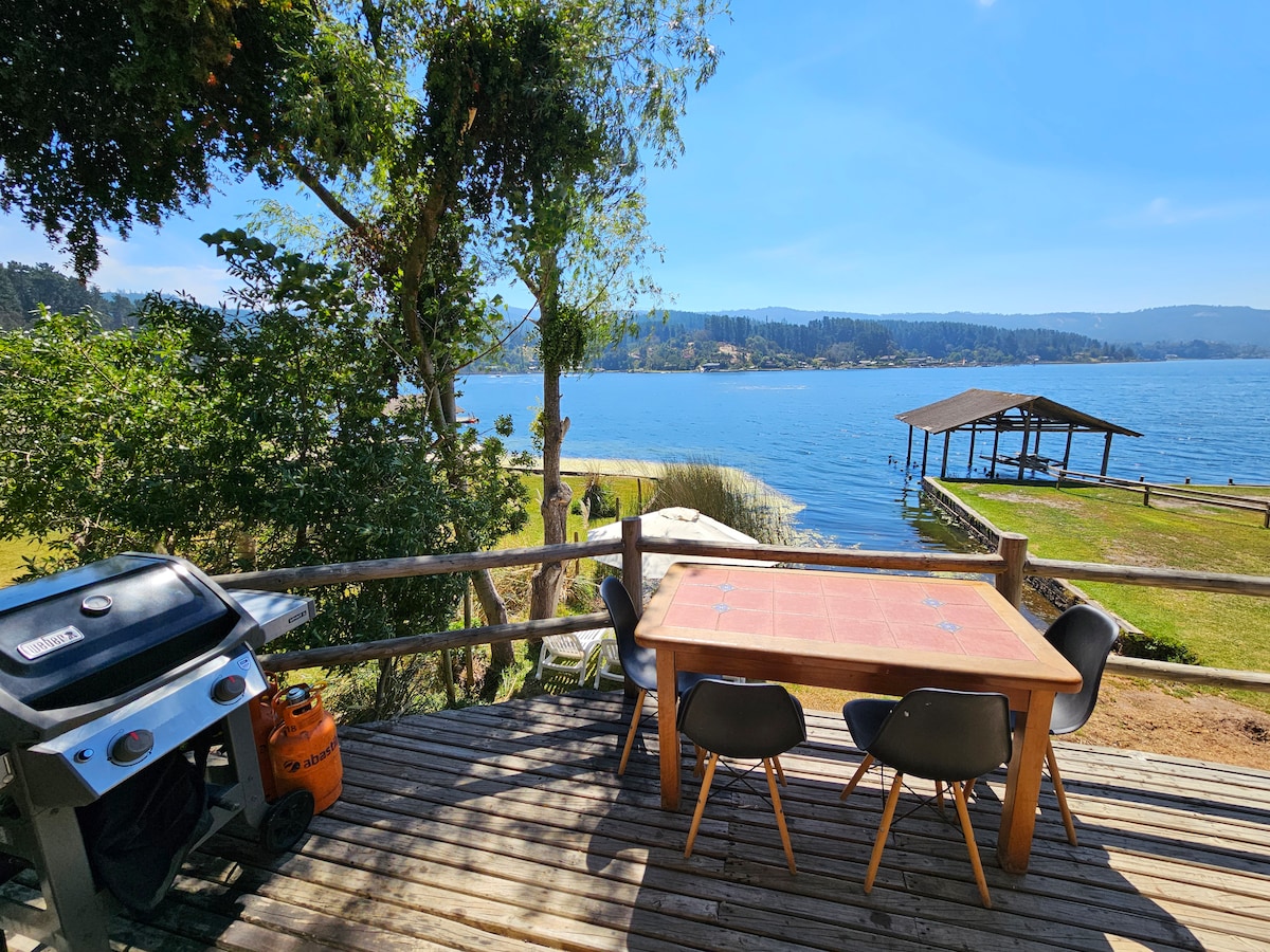 A wooden deck is viewed overlooking a serene lake, with a table and chairs positioned for outdoor dining. A barbecue grill is nearby, and the shoreline, dotted with green vegetation, contributes to the scenic environment. A covered structure can be seen on the shore.