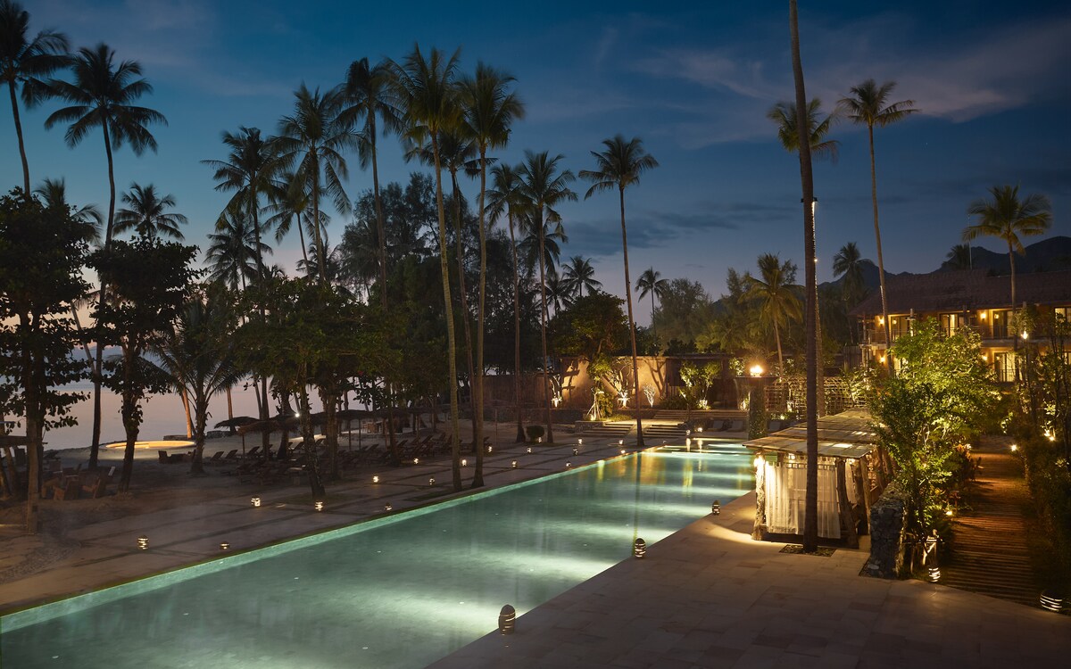An expansive infinity pool is illuminated by soft lighting, framed by tropical palm trees. The serene setting features lounge chairs along the pool's edge, while the ocean can be glimpsed in the background under a twilight sky.