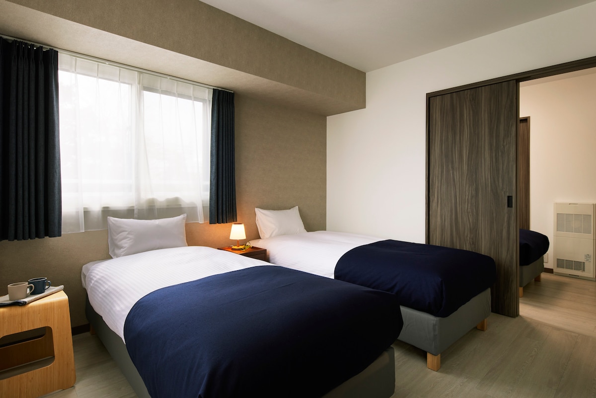A well-appointed guest room features two spacious beds dressed in dark blue linens. Natural light filters in through a large window, complemented by light curtains. A small bedside table holds a lamp, and the room's design incorporates earthy tones for a calming effect.