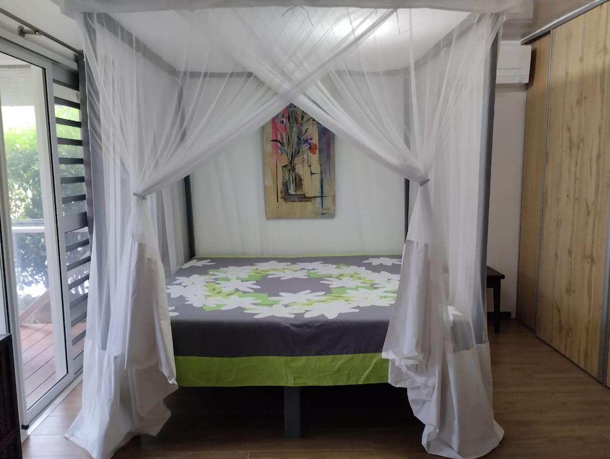 A spacious bedroom features a king-size bed surrounded by a light, flowing canopy. Brightly colored floral artwork hangs on the wall, complemented by natural light filtering through a bay window. A wooden cabinet is positioned nearby, enhancing the room’s cozy ambiance.