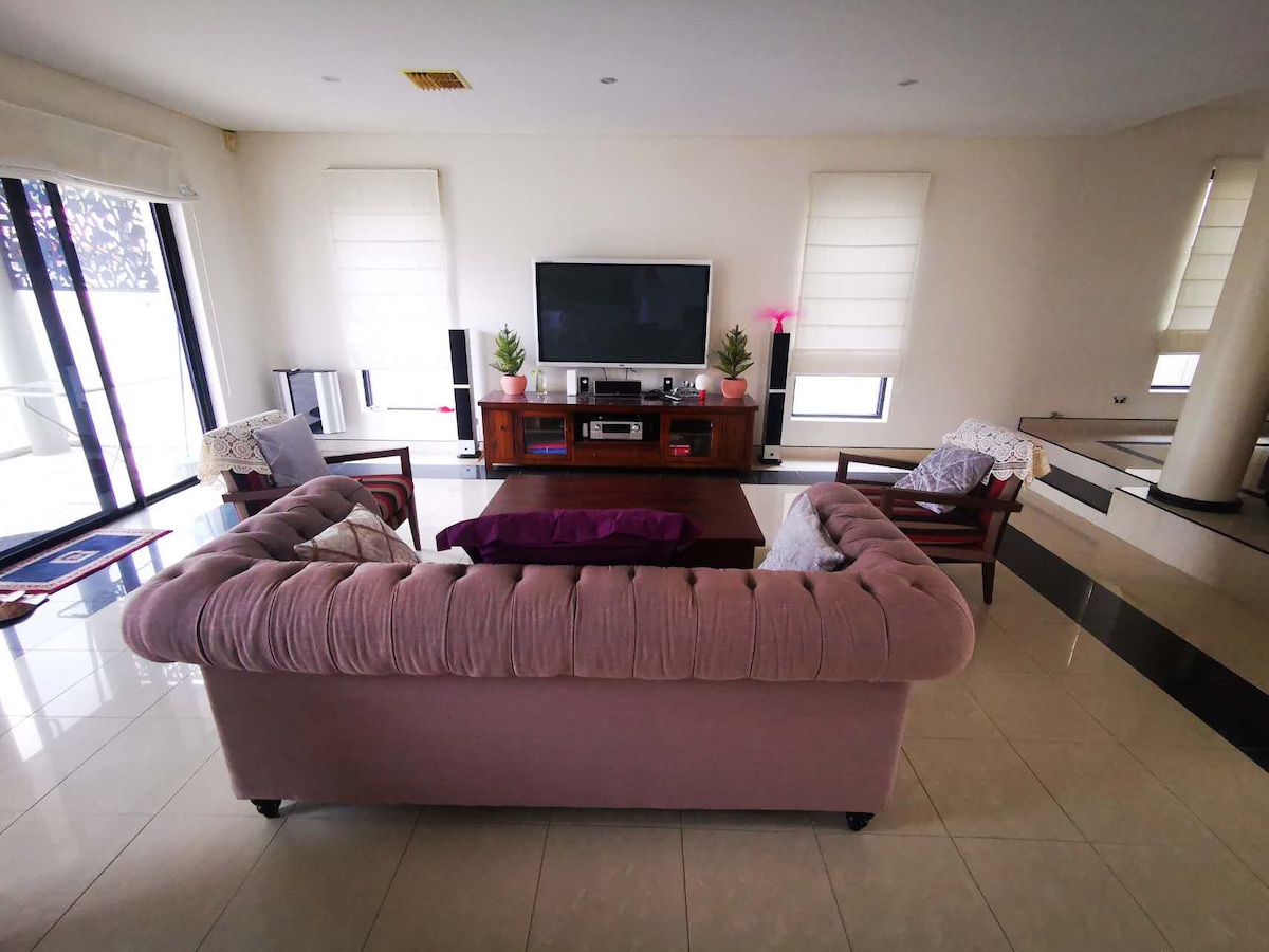 A spacious living room is highlighted by a plush, light-colored sofa and a wooden entertainment unit with a TV. Soft, ambient light enters through large windows adorned with blinds. A low coffee table sits at the center, surrounded by several comfortable chairs.