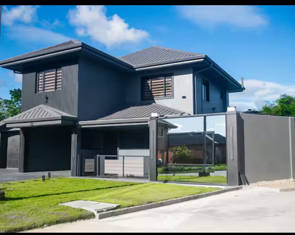 A modern two-story house is depicted, featuring a sleek exterior with a combination of dark materials. Large windows allow natural light to illuminate the interior. A gated entrance is visible, leading to a neatly landscaped front yard with well-maintained grass.