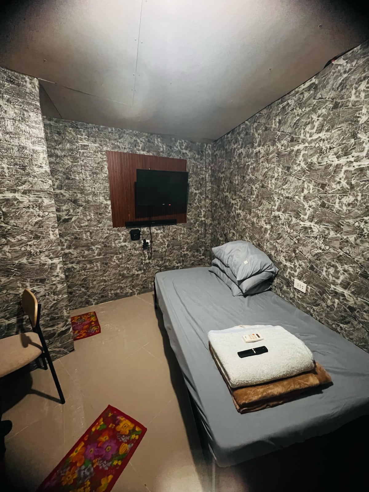 A cozy room is presented with modern wallpaper in subtle patterns. A neatly made bed is equipped with a plain gray blanket and a towel placed on top. A small television is mounted on the wall, and a chair is positioned nearby on the light-colored floor.