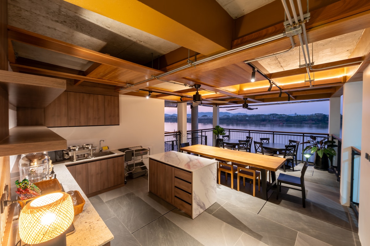 An open-concept kitchen and dining area features modern wooden cabinetry and a spacious marble-topped island. A large dining table is centered with several seats, while large glass doors provide views overlooking the waterfront. Soft lighting enhances the warm tone of the wood accents.