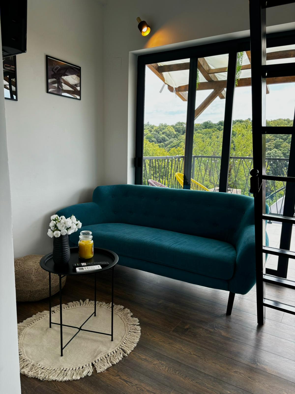 A cozy seating area is highlighted by a teal sofa and a round side table with a glass of juice. A small vase with flowers adds a touch of nature. Large glass doors frame the lush outdoor scenery, enhancing the peaceful ambiance.