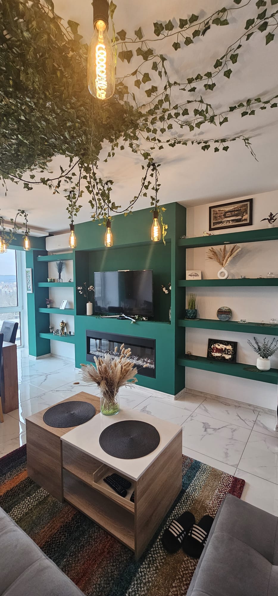 A living room features a green accent wall with built-in shelves displaying decorative items. A flat-screen TV and an electric fireplace are positioned centrally. Hanging lights illuminate the space, while a coffee table with coasters sits on a colorful area rug. Potted plants add a natural touch.