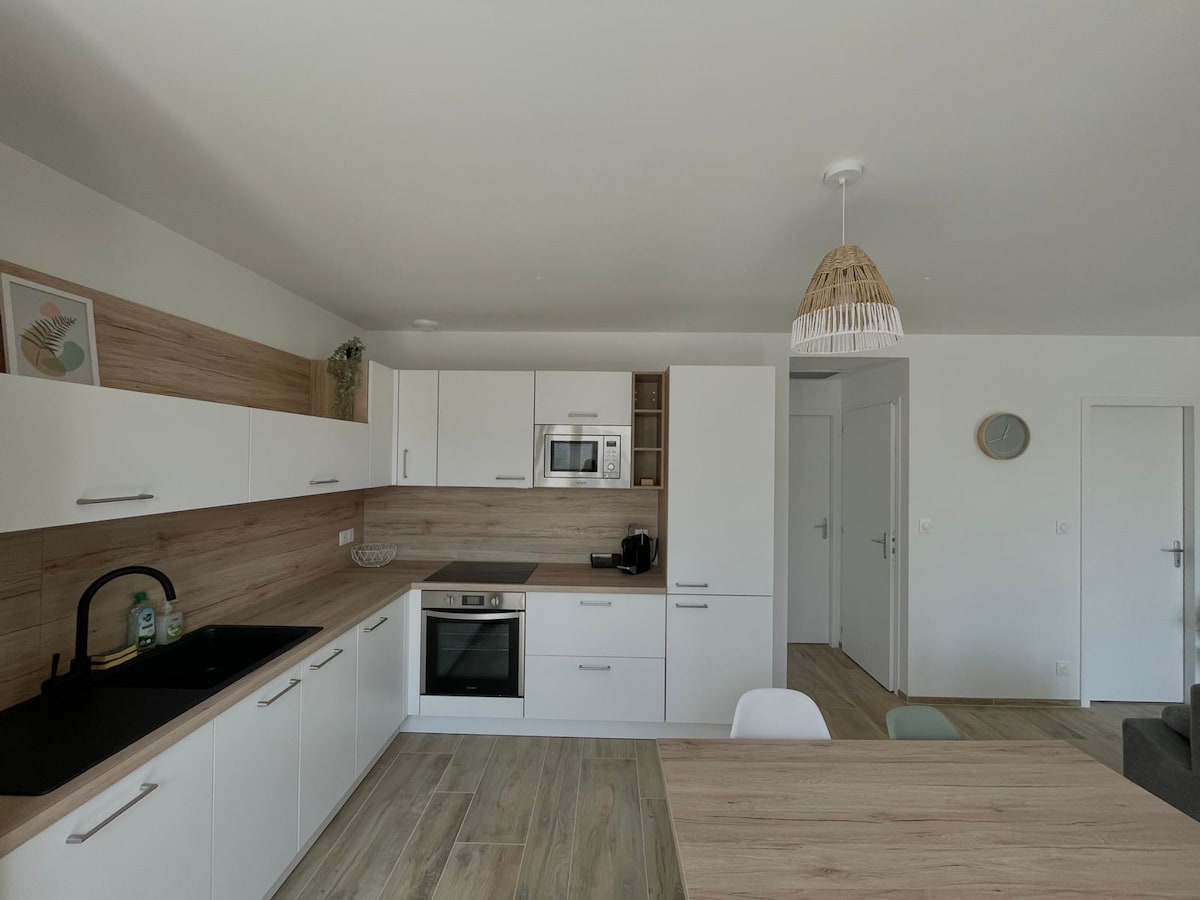 A modern kitchen features sleek white cabinetry complemented by wooden accents. Built-in appliances, including an oven and microwave, are visible. A spacious countertop provides ample preparation space, and a dining area is seen in the foreground. Natural light enhances the airy atmosphere, with decor items adding warmth.