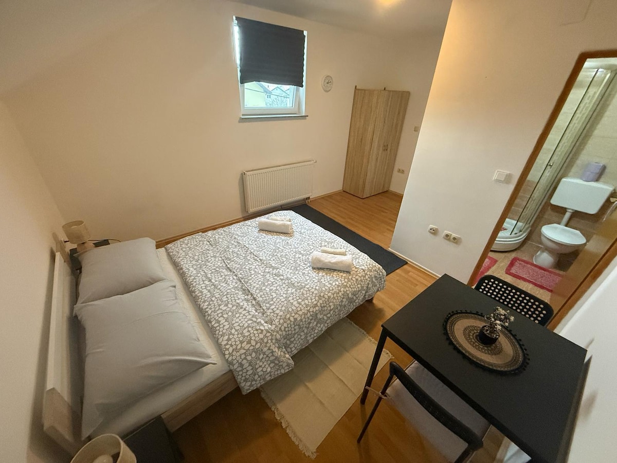 A clean and simple bedroom is showcased with light wooden flooring. A spacious bed is adorned with gray linens and two folded towels. A small table and chair are positioned next to the bed, while a wardrobe is visible in the background. Natural light enters through a window covered by a blind.
