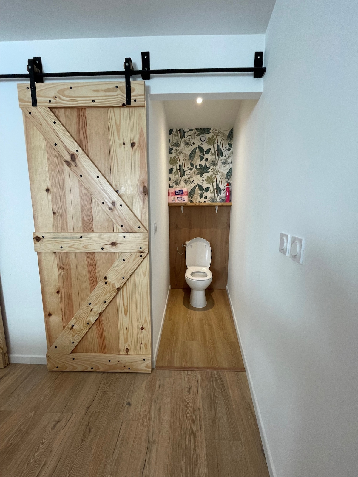 A rustic wooden sliding door leads to a compact restroom. The interior features light wood paneling and a wall adorned with botanical wallpaper. A white toilet is positioned against the back wall, with soft lighting enhancing the clean and tidy atmosphere.