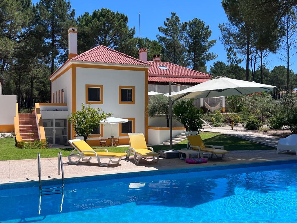 A charming building with yellow accents is viewed beside a clear swimming pool. Sun loungers are arranged next to the pool, with umbrellas providing shade. Lush greenery surrounds the area, creating a serene outdoor environment ideal for relaxation.