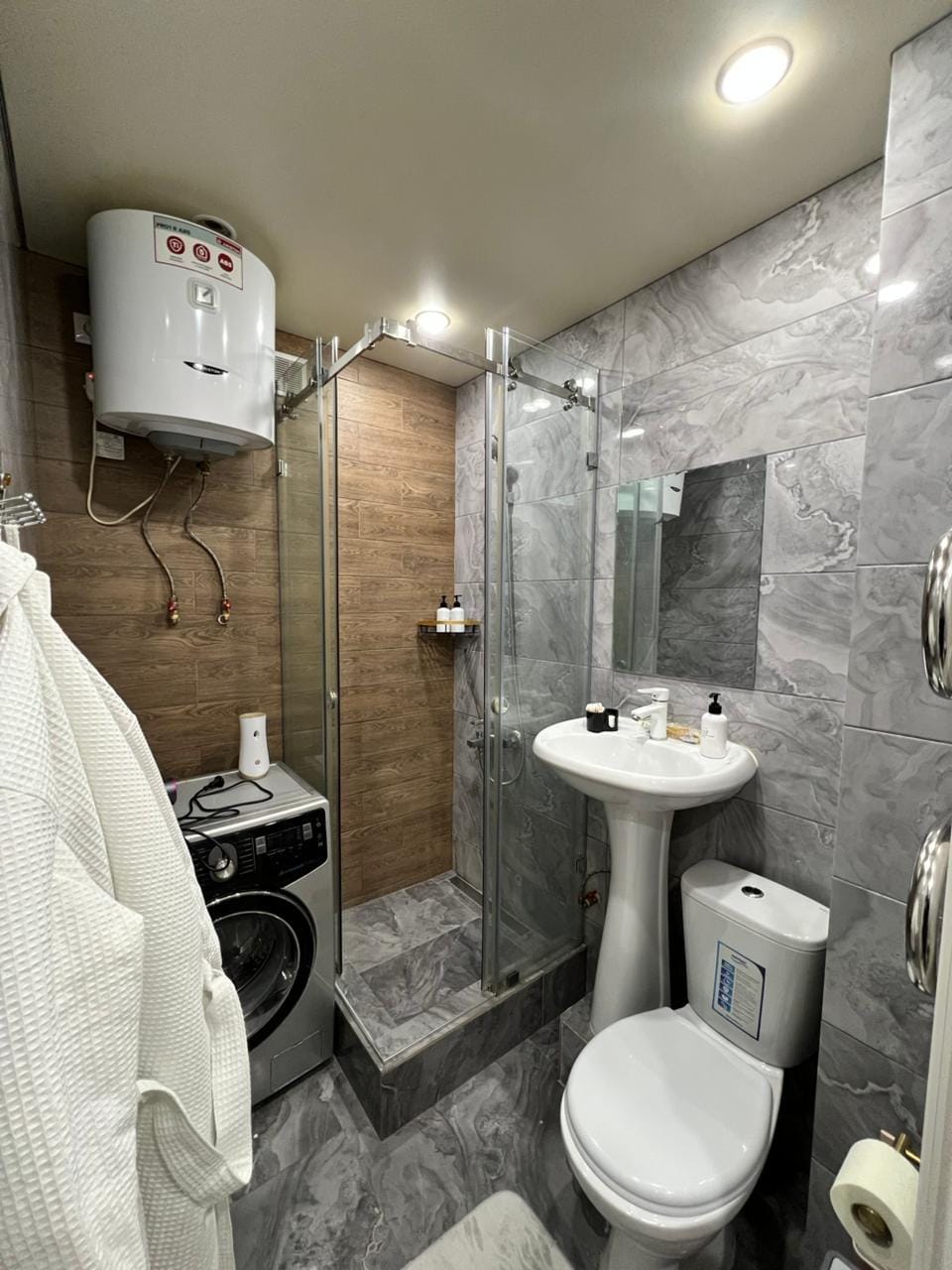 A modern bathroom is presented, featuring a glass shower enclosure and a sleek vanity with a mirror above. There is a washing machine positioned nearby. Soft towels are hung, and toiletries are organized on the countertop.