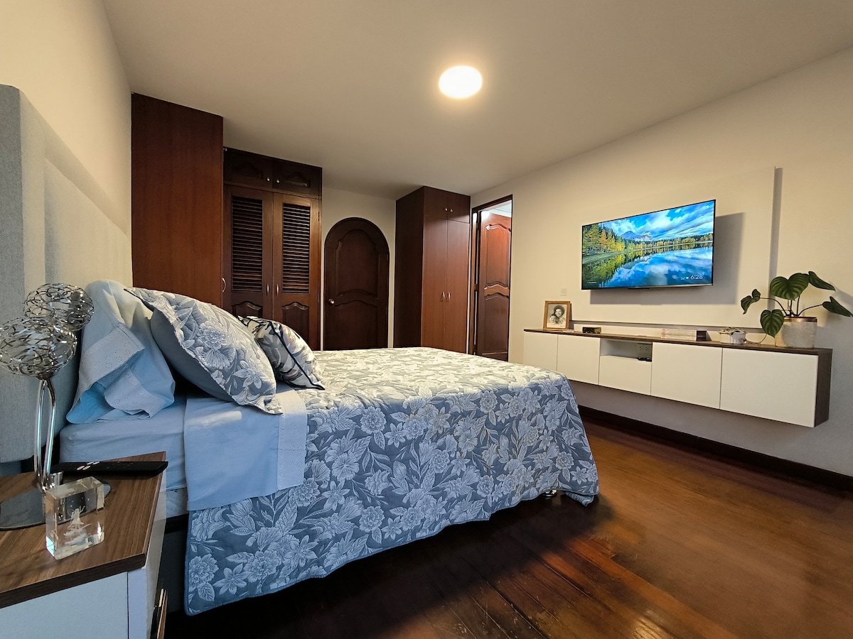 A spacious room is furnished with a comfortable bed covered in a floral-patterned blanket. A large smart TV is mounted on the wall across from the bed, while wooden furniture adds a warm touch. Ample closet space is visible in the background.