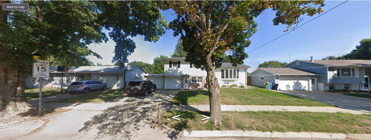 A residential neighborhood scene is captured, featuring a two-story home with large windows. The front yard is bordered by trees, providing shade. Driveways are visible, accommodating vehicles. A street sign indicating a speed limit of 15 mph is present, contributing to the peaceful atmosphere of the area.