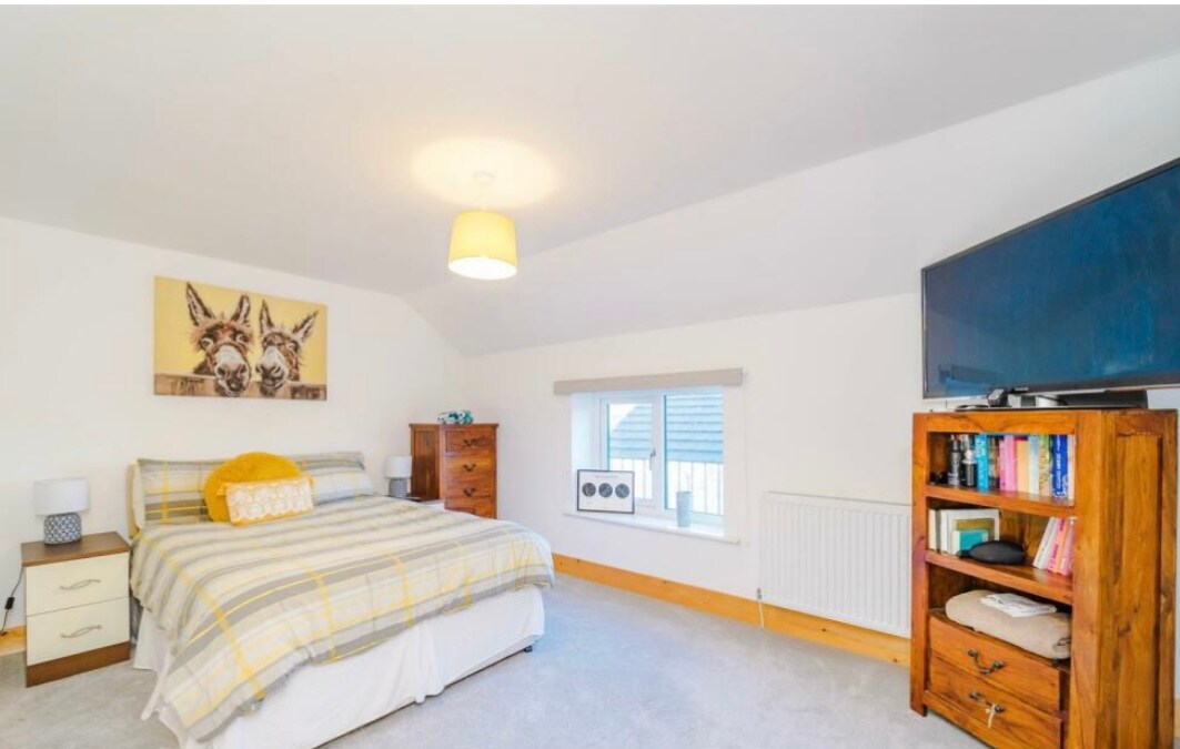 A bright bedroom is presented, featuring a spacious bed dressed in a striped blanket. Two bedside lamps are positioned next to the bed, and a wooden dresser is placed against the wall. A flat-screen TV is mounted nearby, with a window allowing natural light to fill the room.