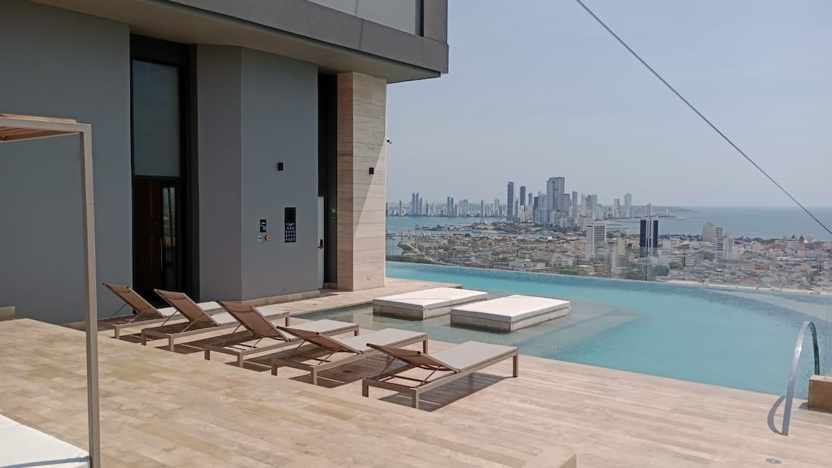 An expansive terrace features lounge chairs and sun loungers overlooking a stunning panoramic view of Cartagena. A sleek infinity pool extends towards the edge, blending seamlessly with the horizon, offering a serene space for relaxation amidst the urban skyline and Caribbean Sea.