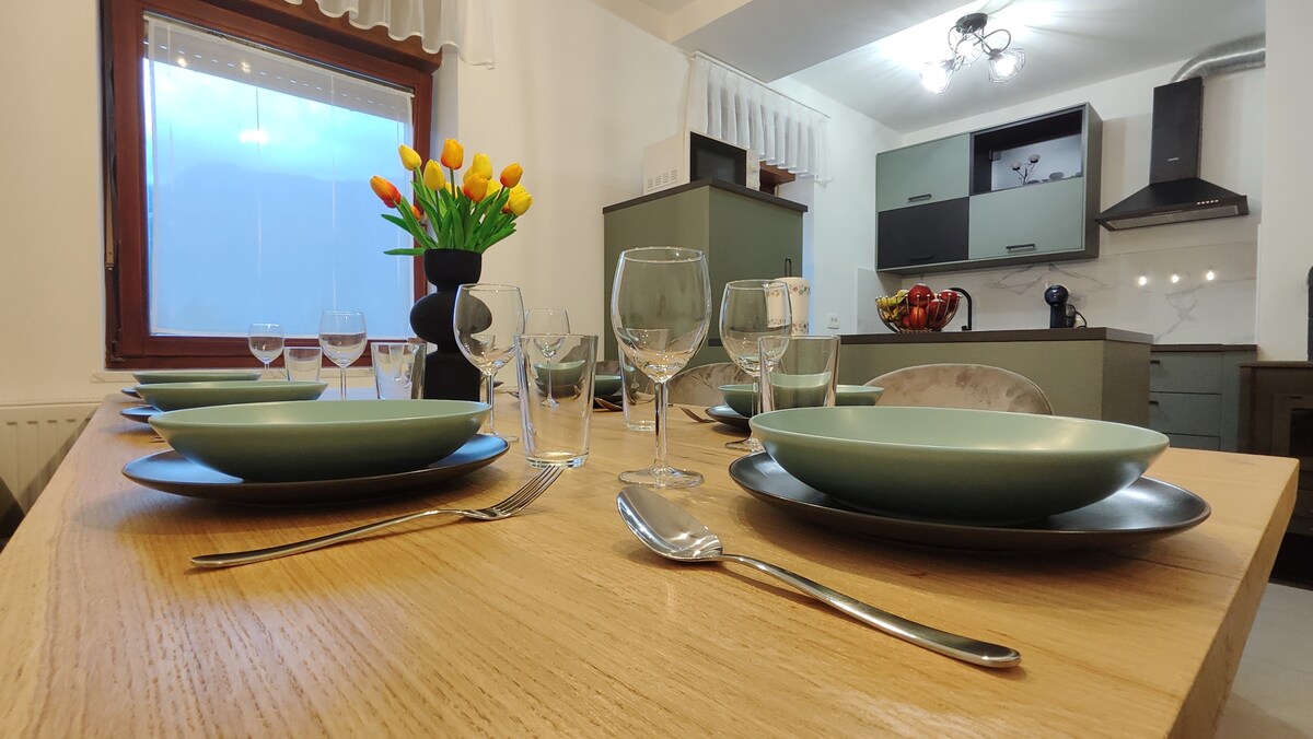 A dining table is set with green plates and polished cutlery. Clear glassware is positioned alongside, reflecting light from the nearby window. A vase with vibrant tulips adds a touch of color, while a modern kitchen area can be seen in the background.