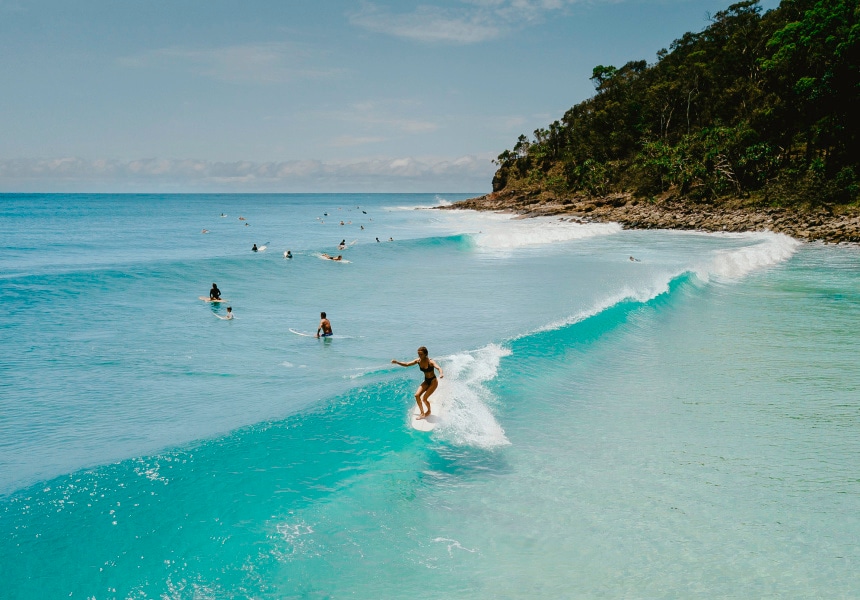 A clear view of surfers riding waves in turquoise water, with a calm beach setting and lush greenery nearby. The horizon is visible with a few clouds, contributing to a serene coastal atmosphere.