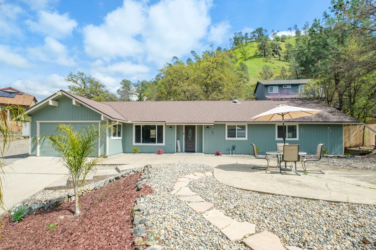 A single-story home is set against a lush hillside, featuring a large paved patio area. A stone pathway leads to the entrance, surrounded by gravel landscaping and colorful plants. A patio table with chairs and an umbrella is positioned for outdoor dining.