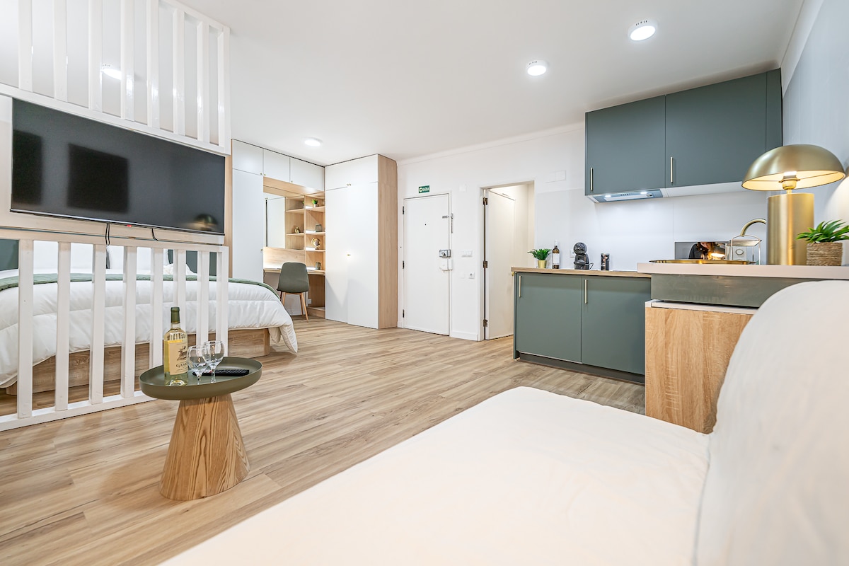 A modern studio layout showcases a living area with a neutral sofa and a stylish circular table. A wall-mounted HDTV is visible, alongside a fully equipped kitchen with contemporary cabinetry. The workspace is conveniently positioned, and the entrance door is clearly identified.