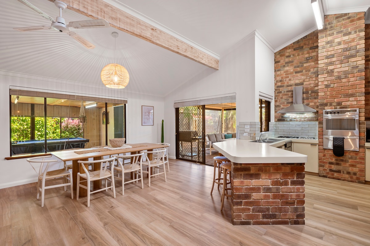 A bright kitchen and dining area is showcased, featuring a large dining table with white chairs and a modern kitchen countertop. Natural light pours in through wide windows, highlighting brick accents and a neutral color palette. A ceiling fan and pendant light provide added comfort and style.